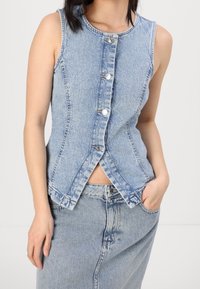 Žena v bezrukávové, zapínací svetříku ze světlého modrého denimu a odpovídající sukni s vysokým pasem, má ruku v kapse.