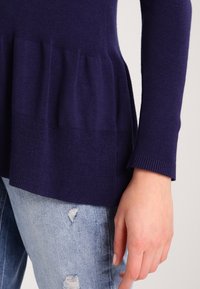 Un haut tricoté bleu marine avec un design texturé à volants à la taille et des manches longues, accompagné de jeans déchirés bleu clair.