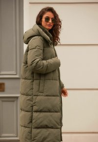 Manteau matelassé long de couleur vert olive avec capuche, motif matelassé et fermeture éclair sur toute sa longueur. Le mannequin porte des lunettes de soleil rondes et se tient contre un fond neutre.