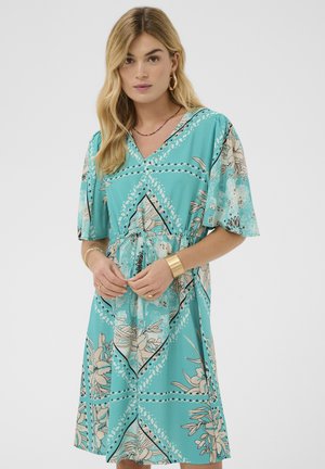 Femeie purtând o rochie până la genunchi cu flori în culoarea teal, cu mâneci scurte, brățară aurie și cercei rotunzi, ajustând șnurul de la talie pe un fundal simplu.