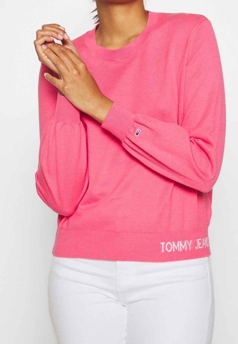 Femme portant un pull à manches longues rose vif avec le texte « TOMMY JEANS » près de l'ourlet et un pantalon blanc, sur un fond uni.