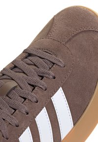 Sneaker in suede marrone con punta arrotondata, lacci piatti, strisce bianche sui lati e suola in gomma naturale per una buona aderenza.