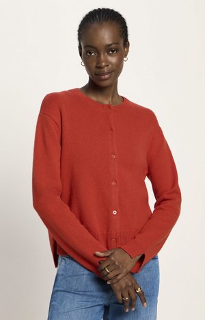 Woman wearing a red buttoned sweater and blue jeans, with hands crossed, wearing gold rings and small hoop earrings, neutral background.