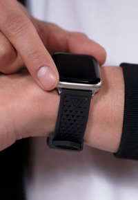 Smartklocka med ett svart gummiband som har ett texturerat triangulärt mönster, silverfärgat rostfritt stålfodral och en slät svart skärm.