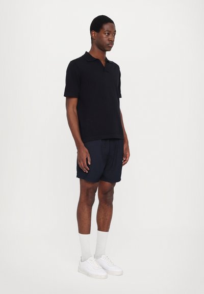 Polo preto de manga curta, calções azul-marinho, sapatilhas brancas e meias claras. Design simples com um corte reto e textura suave.
