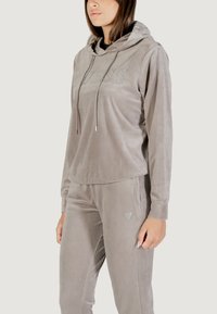 Chándal de terciopelo gris que incluye una sudadera con capucha con la palabra "GUESS" en pedrería y pantalones a juego con un pequeño logo en el lateral.