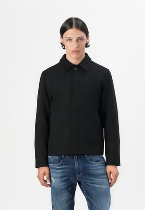 Black outer jacket with a collar, single button closure, and side pockets. Worn over blue denim jeans. Simple design and structured shape.