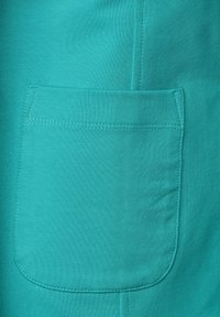 Tissu turquoise à texture lisse avec une poche rectangulaire cousue sur le devant. Des coutures décoratives entourent la zone de la poche.