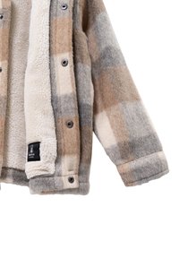 Giacca a quadri in beige, grigio e crema. Interno in felpa, bottoni a pressione e texture morbida. Etichetta visibile con la scritta "CALDO", che indica calore.