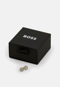 Srebrni manšeti z okroglo obliko in teksturirano površino ob črni škatli z belim logotipom "BOSS" in zanko za vlečenje.