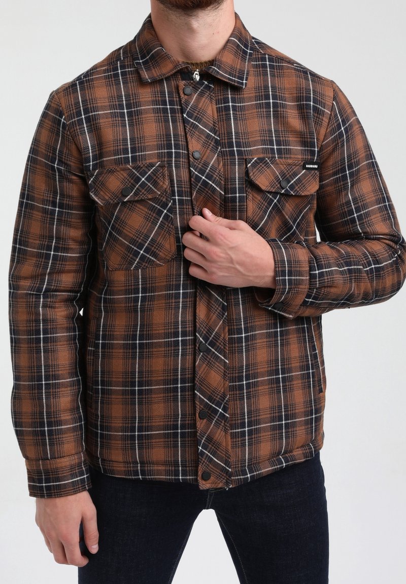 Homme portant une chemise à carreaux marron et noire à manches longues avec des poches poitrine boutonnées, en train d'ajuster les boutons à l'avant, associée à un jean foncé.