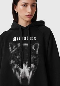 Sudadera con capucha negra con un gran gráfico de un lobo en la parte frontal, con el texto en negrita "Allsaints" encima, y cordones negros con puntas de plata.