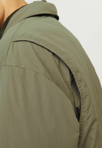 Olive green lightweight jacket featuring a relaxed fit, textured fabric, and pronounced shoulder seams, emphasizing a contemporary design.