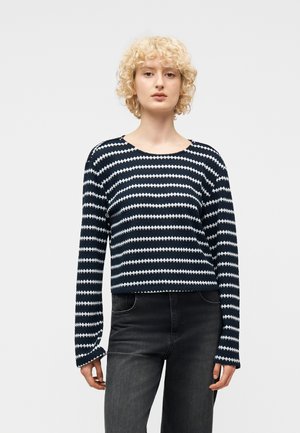 Giovane con capelli biondi ricci che indossa un maglione a maniche lunghe a righe blu navy e bianche e jeans grigio scuro, in piedi davanti a uno sfondo chiaro e semplice.