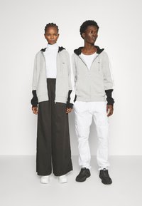 Felpe grigie con zip e dettagli neri, indossate sopra un dolcevita bianco e una camicia bianca. Pantaloni larghi e scuri; scarpe ginniche bianco e nere.