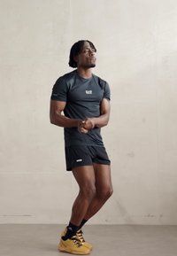 T-shirt sportivo nero con logo EA7, abbinato a pantaloni corti neri e scarpe da ginnastica gialle. Il modello si trova su uno sfondo neutro.