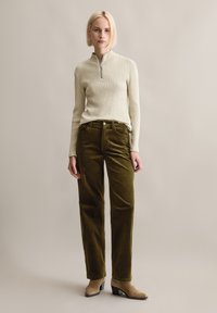 Rippstrickcreme-Pullover mit einem halben Reißverschluss, kombiniert mit olivgrünen Cordhosen. Das Modell trägt beigefarbene Ankle-Boots vor einem schlichten Hintergrund.