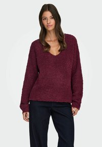 Burgunderfarbener, flauschiger Pullover mit tiefem V-Ausschnitt, langen Ärmeln und lockerer Passform, kombiniert mit dunklen Jeans. Weiche Textur und gerippte Bündchen.