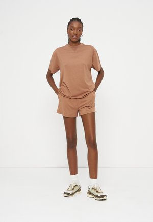 Žena stoji s rukama u džepovima, nosi smeđu košulju kratkih rukava i kratke hlače, bijele čarape i bež tenisice na bijeloj pozadini.