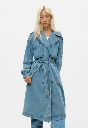 Kobieta o długich blond włosach, ubrana w przewiązany paskiem, dwurzędowy płaszcz trencz z denimu oraz dopasowane spodnie z denimu na białym tle.