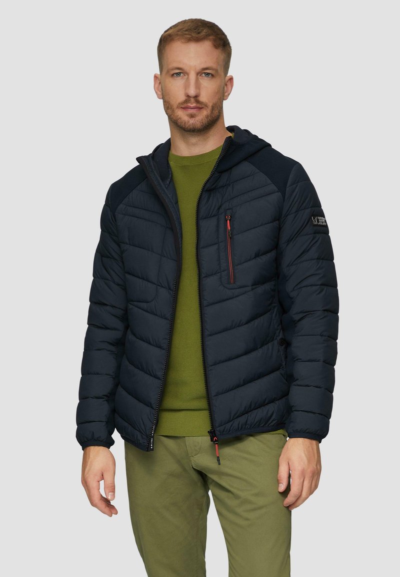 Marineblauwe pufferjack met capuchon, geborduurd ontwerp, met een zijzak met rits en contrasterende donkere stofaccenten, gecombineerd met een groene trui.