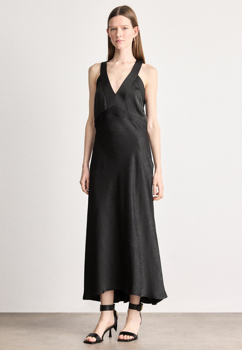 Čierne saténové maxi šaty s hlbokým výstrihom do V, širokými ramienkami a voľným siluetou. Skombinované s čiernymi sandálmi na členkovom opätku.