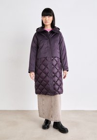 Cămașă căptușită de culoare violet, cu o jumătate inferioară matlasată, având închidere cu fermoar, glugă și un material superior neted. Puritată peste o fustă ușoară și cizme.