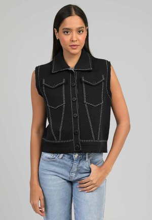 Femme aux cheveux noirs lisses portant un gilet noir sans manches boutonné avec des coutures blanches et un jean bleu clair, debout devant un fond gris.