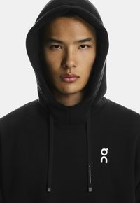 Juodas gobtuvėlis su balta logotipu ant krūtinės, reguliuojamais raišteliais ir minkšta, tekstūruota medžiaga.