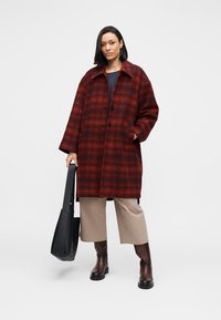 Red and black checkered wool coat with a collar, button closure, and side pockets, worn over a navy top and beige wide-leg pants.