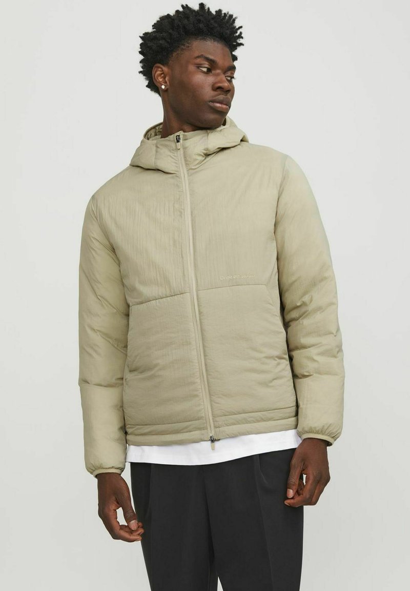 Doudoune légère beige avec capuche, en tissu texturé, fermeture éclair à l'avant et deux poches horizontales. Porté par-dessus une chemise blanche.
