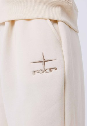 Beige sweatpants van zacht materiaal, met een geribbelde tailleband en zijzakken. Een geborduurd "PXP" logo is zichtbaar op de rechterzak.