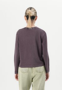 Persoon met blond haar in een knot, draagt een donkerpaarse longsleeve en lichtgroene spijkerbroek, staande voor een effen witte achtergrond.