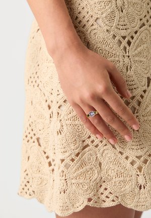 Anillo de oro con una gema púrpura rectangular, llevado en una mano contra un vestido de encaje crema con crochet que presenta patrones y texturas intrincadas.
