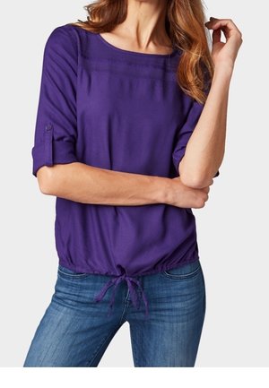 Kobieta w fioletowej bluzce z długim rękawem, z wiązaniem u dołu i podwiniętymi rękawami, w połączeniu z niebieskimi dżinsami, stoi z założonymi rękami.