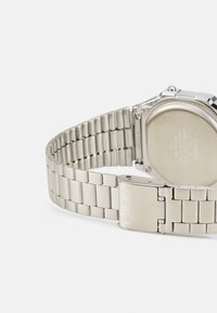 Montre en acier inoxydable avec un bracelet en métal argenté, boîtier carré et un fermoir arborant le logo "CASIO". Des maillons lisses et texturés sont visibles.