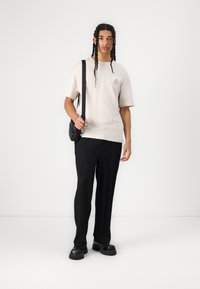 Beige bomulds T-shirt med broderi på brystet, parret med sorte ribbede bukser og rundede sorte sko. Mand holder en lille sort taske.