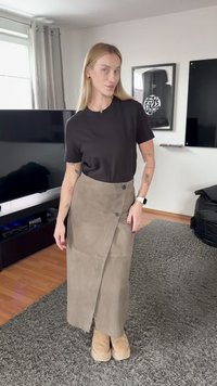 Kvinde med langt blondt hår iført en sort t-shirt, beige omslagsnederdel og beige sko, der står på et gråt tæppe i en stue.