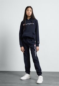 Ensemble sweat à capuche marine et pantalon de survêtement en mélange de coton, avec le logo Champion blanc, coupe décontractée et poches latérales. Associé à des baskets blanches.