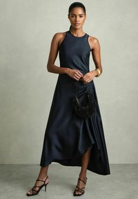 Sort satin kjole med høj halsudskæring, asymmetrisk kant og draperede detaljer. Komplementeret af en tekstureret sort håndtaske og strappy højhælede sko.