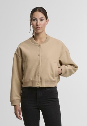 Tricou scurt bej cu guler rotund, nasturi în față și mâneci mari. Include un buzunar lateral și tiv și mansete ribbate.