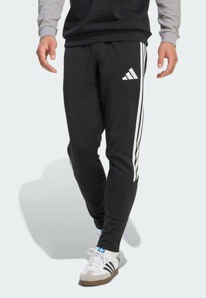 Person iført sorte Adidas træningsbukser med hvide striber og Adidas-logo, gående, kombineret med hvide Adidas sneakers og en lysegrå sweatshirt.