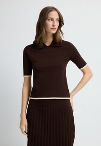 Brun polo-stil top med korte ærmer, cremefarvet kant ved krave og nederste kant, glat tekstur, tætsiddende design. Båret med plisseret brun nederdel.