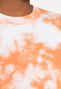 Sudadera de cuello redondo de manga corta, blanca con patrón de tie-dye naranja, material de algodón, superficie texturizada, costuras visibles alrededor del cuello.