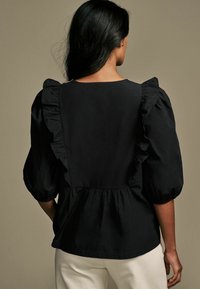 Blusa nera con maniche a sbuffo e dettagli a volant, vita cintrata e semplice scollo rotondo. Tessuto liscio, design minimalista.