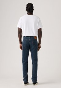 Jeans de ganga lavagem escura com corte de perna reta, remendo de couro castanho nas costas e bolsos traseiros. Combinados com uma t-shirt branca simples.