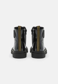 Bottines noires en cuir verni avec une semelle épaisse, des rayures latérales texturées jaunes et un emblème rond à l'arrière.