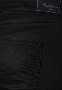 Jeans de mezclilla negros con un bolsillo trasero que presenta un diseño de costura ondulada y una etiqueta de cuero con marca en la cintura. Textura suave.