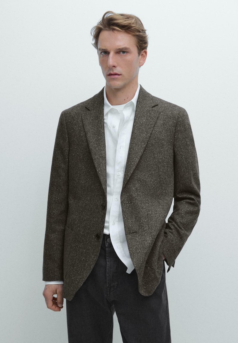 Brauner Tweed-Blazer mit strukturiertem Finish, einreihiger Schnitt, zwei Knöpfe, Reverskragen, kombiniert mit einem weißen Hemd und dunkler Jeans.