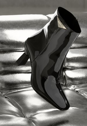 Botte de cheville en cuir verni noir avec un bout pointu et un talon carré. Équipée d'une plaque logo dorée et d'une finition lisse et brillante.